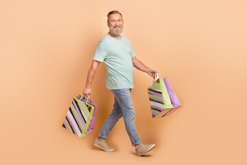 Full length profile photo of cheerful person hold shop bags walking isolated on beige color background
