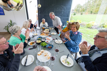 A group of seniors enjoys a convivial meal together around a dining table filled with an assortment of dishes and wine. A man stands, possibly giving a toast or sharing a story, as he is greeted with
