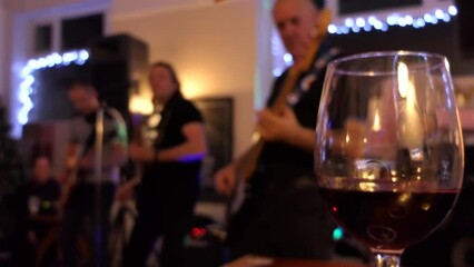 Close up shot of glass of wine, blurry view of band playing and singing live in the pub, three guitarists and a drummer, obstructed by glass; selective focus.