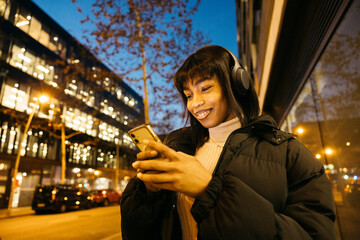Young woman walking with wireless headphones and using a phone while walking on a city.