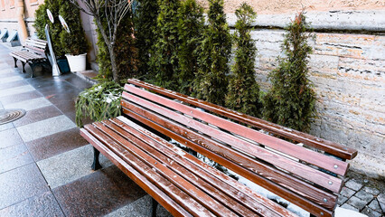 Wooden bench on wet pavement outdoors. Spring in city. Winter in town. Urban landscape with fir...