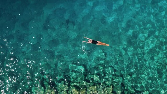 Top down view of young woman in black swimsuit floating on water surface in crystal clear turquoise ocean. Happy girl on vacation at paradise.