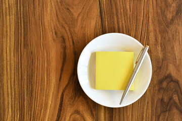 Libreta de notas y una pluma en un plato blanco sobre una mesa de madera, espacio para texto al lado izquierdo.