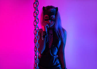 Sexy girl in latex BDSM mistress dress and cat mask in neon light on dark background with chains for sex game