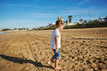 Adorable Caucasian blonde child girl in white t shirt and blue jeans, walking barefoot on the wet sand