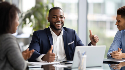 A confident professional man in a suit is giving a thumbs up in a bright office environment with cheerful colleagues around him, indicating success and approval.