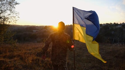 Young man in military uniform jogs with waving flag of Ukraine at sunset. Male soldier of ukrainian...