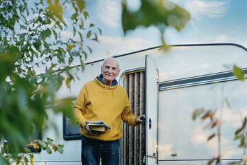 Retired man with book and newspaper in hands standing in door of his motor home outside an rv