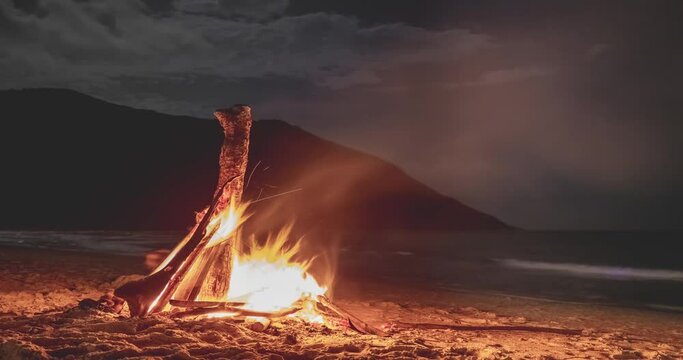 Beach bonfire at night under moonlight. Timelapse above mountain island. Burning wood on sand shore. Dark sky with stars. Tropical romantic landscape near ocean with waves. Camp atmosphere.