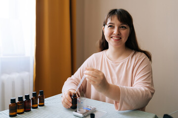 Cheerful female craftswoman preparing using of scented oil for creating candle building mixture. Process of making handmade natural scented candle at workshop. Concept of creative handicraft.