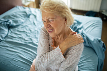 Senior woman in pain holding shoulder in bedroom at home