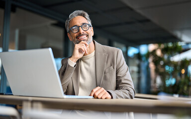 Happy mature business man entrepreneur using laptop computer outside office looking away. Smiling busy middle aged professional male entrepreneur sitting at table outdoors hybrid working. Copy space