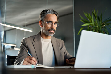 Mature busy businessman entrepreneur or executive writing notes elearning webinar sitting at desk,...
