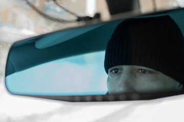 The rearview mirror reflects the concentrated eyes of the driver in a winter hat in the car interior