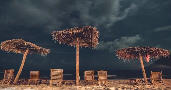 Empty hotel lounge chairs and tiki umbrellas on beach as sunset glows in sky, Timelapse. Beach umbrellas at night. Koh Phangan. Thailand
