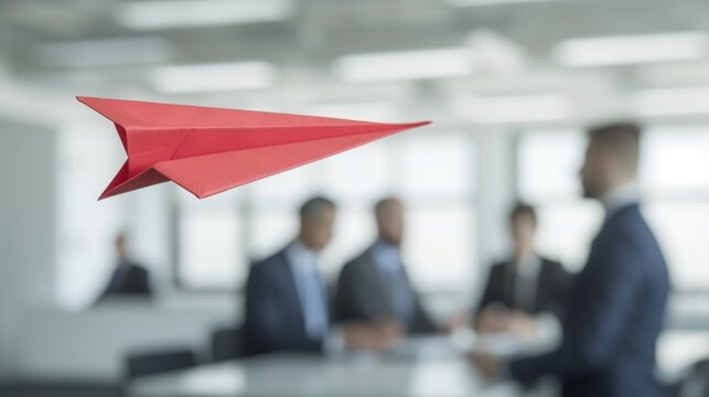Red paper plane flying in an office with people in the background