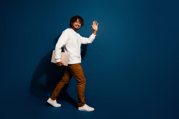 Fototapeta na wymiar Full length photo of good mood funky guy dressed white pullover holding modern device waving hi empty space isolated dark blue color background