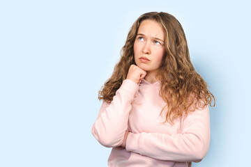 Young cute thoughtful intelligent woman with curly hair looking up, hand on chin isolated over blue background, copy space, deep thinking, creative person