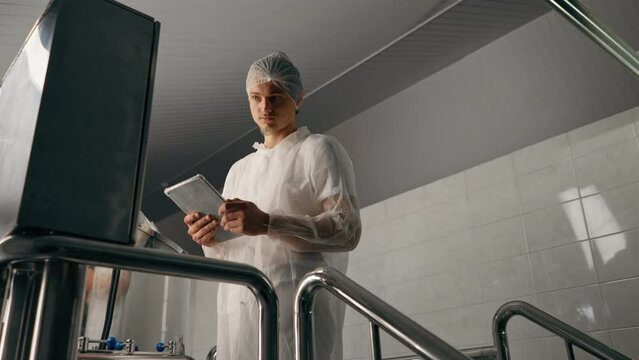 A male technologist in a protective suit with a tablet in his hands checks the production stage of the fermentation of a drink at a brewery