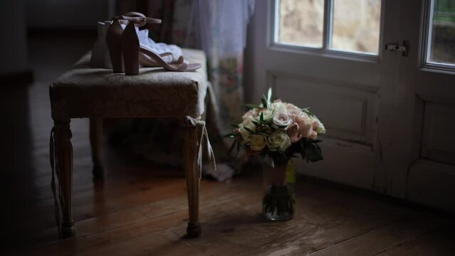 Imagen est&aacute;tica de zapatos de novia sobre una silla, un ramo de flores en un florero en el suelo frente a un ventanal