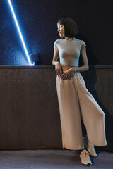 Stylish woman in wide trousers and a T-shirt poses at night in the city. - 738241696
