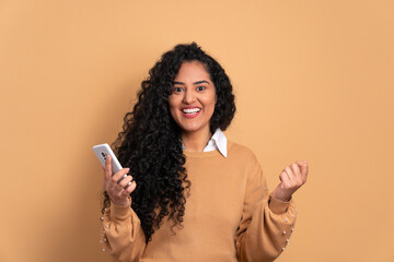 excited black young woman smiling and holding mobile phone in beige studio background. joy, people concept.