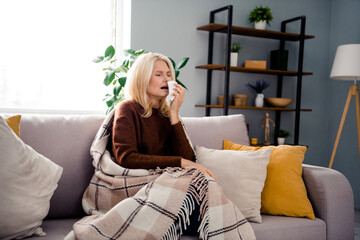 Photo of mature age grandmother sitting couch sneezes using napkin illness at home need healthcare...