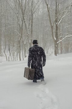Retro Man walking away in snow carrying vintage suitase wearing long coat boler hat