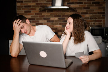 Married couple sits in kitchen at a laptop and analyzes their finances and debts. Man is shocked by...