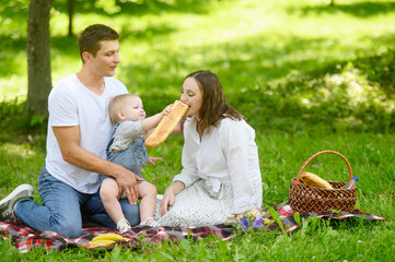 Dad, mom and daughter are sitting in a meadow on a blanket in the park on a warm sunny day,child holds a baguette in his hands, treats his mother. Family weekend getaway