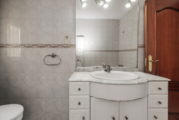 a bathroom with white tiled wall, white wooden cabinet with porcelain sink, frameless glass mirror...