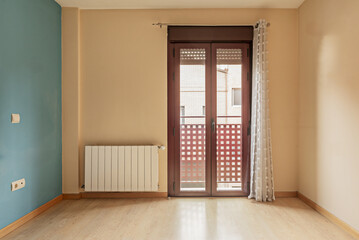 An empty room with a blue painted wall, a balcony with brown aluminum and glass double doors and a white aluminum radiator