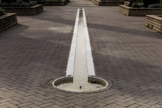 A small circular fountain at ground level with a small elongated pond filled with water on the bricked floor of an urban park