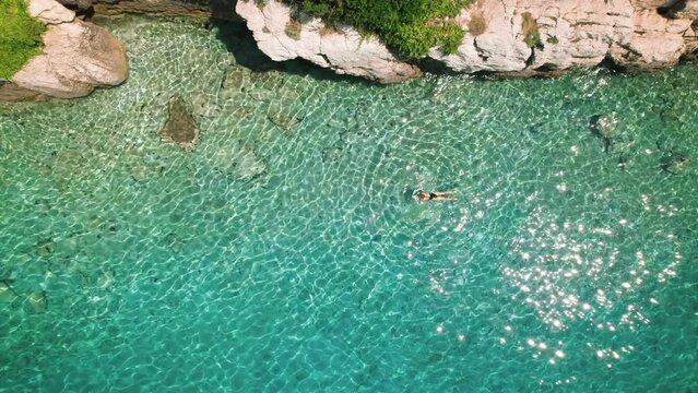 A lone swimmer woman enjoys the crystal-clear waters near a rocky shore. Aerial view of serene ocean expanse.