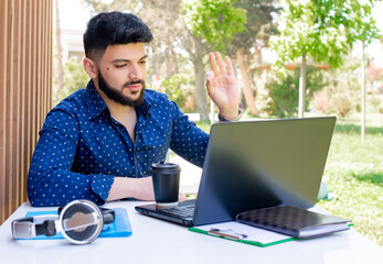 Front view of brunette young male with beard sititng at table, using laptop. Asian businessman working, looking at screen, raising hand, vawing. Concept of urban lifestyle.