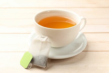 Tea bag and cup of hot beverage on light wooden table, closeup