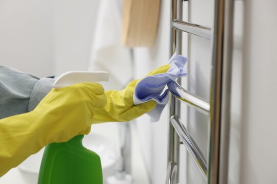 Woman cleaning heated towel rail with sprayer and rag, closeup