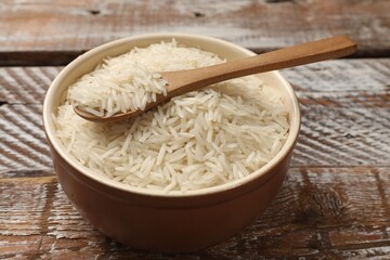 Raw basmati rice and spoon in bowl on wooden table, closeup