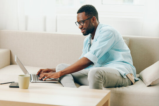 Smiling African American Man Working on Laptop in Cozy Home Office, Talking on Phone and Enjoying Online Shopping This modern and cheerful image depicts a male freelancer sitting on a comfortable sofa