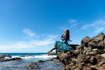 Beautiful woman in blue clothes and braided hair sitting on the edge of the beach rocks sunbathing