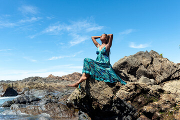 Beautiful woman in blue clothes sitting on the edge of the beach rocks touching her hair braids