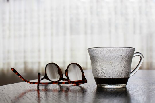 Taza de cristal grabado con un poco de café, junto a unos lentes, frente a una ventana