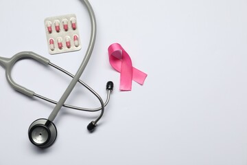 Breast cancer awareness. Pink ribbon, stethoscope and pills on white background, flat lay. Space...