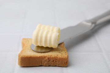 Tasty butter curl, knife and piece of dry bread on white tiled table, closeup