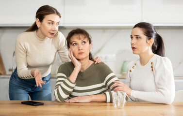 Upset woman in her middle years is seated at table supported by other kind women in the kitchen