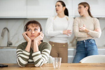 Upset middle-aged woman sitting at the kitchen table with her back to women quarreling angrily to...