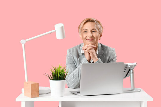 Mature female programmer working with laptop at table on pink background