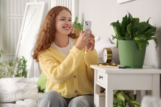 Young woman with mobile phone taking picture of plant in bedroom