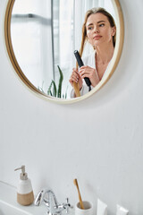 attractive blonde woman in cozy bathrobe using flat iron on her hair and looking at mirror