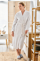 appealing cheerful woman with blonde hair in white bathrobe posing in her bathroom and looking away